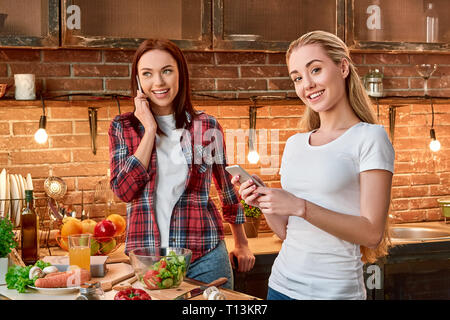 Ritratto di due donne attraenti, divertendosi, durante la preparazione di cibo. Le donne sono sorridente, mentre a parlare e scrivere messaggi tramite telefono. Essi stanno andando a cucinare qualcosa insieme Foto Stock