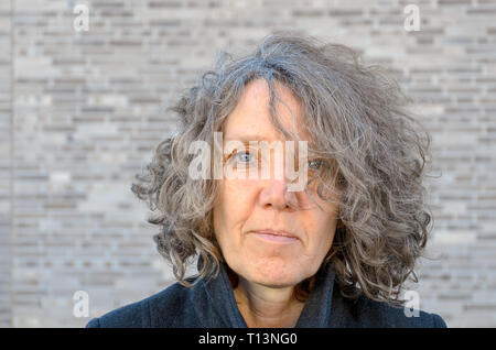 Ritratto di un attraente donna di mezza età con tousled capelli ricci in posa di fronte di grigio di un muro di mattoni guardando nella telecamera Foto Stock