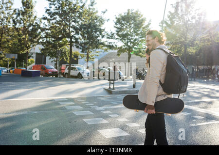 Uomo al telefono con zaino e skateboard attraversando la strada Foto Stock