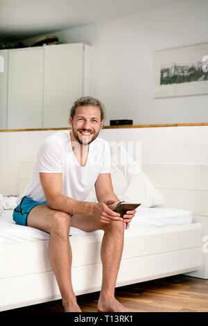 Giovane uomo seduto sul letto, utilizzando digitale compressa al mattino Foto Stock