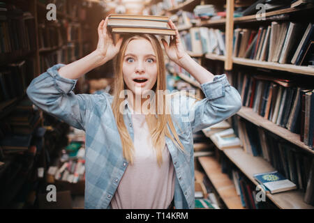 Divertente immagine della ragazza in piedi vicino a scaffale. Lei tiene due libri sulla sua testa con le sue mani e guardando dritto in avanti. Lei si guarda stupito Foto Stock