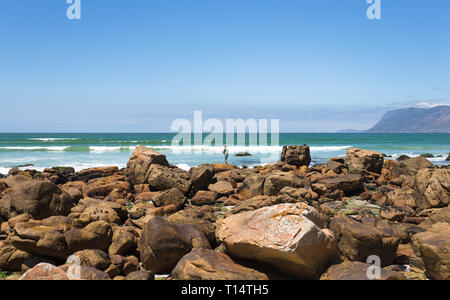 Un solitario mare fisherman sugli scogli lungo la costa con la sua verga e linea sperando di prendere un pesce su una bella giornata estiva con cielo blu Foto Stock