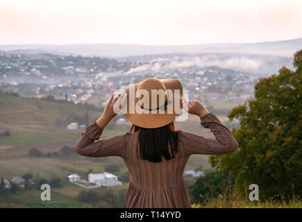 Bella ragazza in un cappello guarda la città al tramonto Foto Stock
