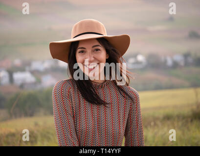 Ritratto di una bella ragazza felice in un cappello su un sfondo della città al tramonto. Gira per la telecamera e dà un bel sorriso giocoso. Foto Stock
