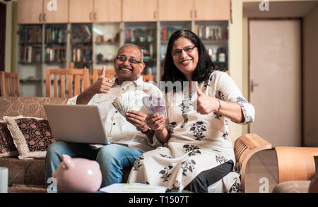 Contabilità di coppia indiana/asiatica senior, facendo finanza domestica e controllando le fatture con il laptop, la calcolatrice e i soldi mentre si siede sul divano/divano a casa Foto Stock