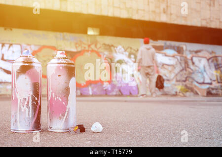 Più usato bombolette spray con rosa e bianco paint giacciono su asfalto contro il ragazzo in piedi di fronte ad una parete dipinta in color disegni graffiti Foto Stock