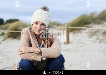 Spagna, Menorca, ritratto di sorridere senior donna che indossa bobble hat sulla spiaggia in inverno Foto Stock