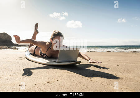 Giovane donna sdraiata sulla tavola da surf, utilizzando laptop Foto Stock