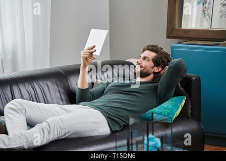 Uomo sorridente con la compressa sdraiato sul lettino a casa Foto Stock