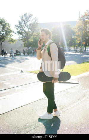 Uomo al telefono con zaino e skateboard in città Foto Stock