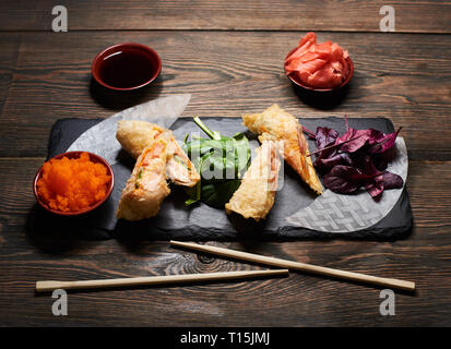 Fritto delizioso sushi rotoli con riso e pesce in nero ardesia servita al tavolo in legno e decorate con bacchette, tazza di salsa di soia di caviale e spro Foto Stock