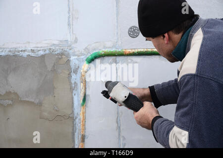 Un anziano operaio trapani un foro in una parete in polistirolo espanso per l'installazione successiva di un rinforzo di plastica il grano. Creazione di fori nel muro con un Foto Stock