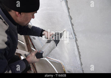 Un anziano operaio trapani un foro in una parete in polistirolo espanso per l'installazione successiva di un rinforzo di plastica il grano. Creazione di fori nel muro con un Foto Stock