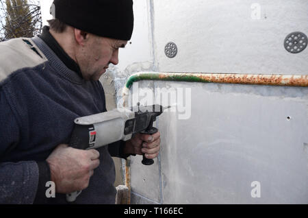 Un anziano operaio trapani un foro in una parete in polistirolo espanso per l'installazione successiva di un rinforzo di plastica il grano. Creazione di fori nel muro con un Foto Stock