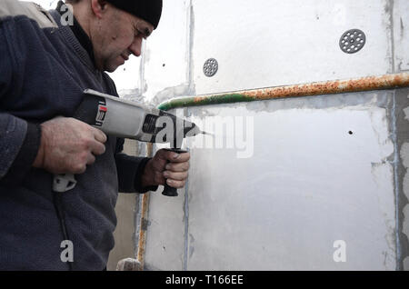 Un anziano operaio trapani un foro in una parete in polistirolo espanso per l'installazione successiva di un rinforzo di plastica il grano. Creazione di fori nel muro con un Foto Stock