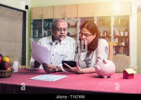 Contabilità di coppia indiana/asiatica senior, facendo finanza domestica e controllando le fatture con il laptop, la calcolatrice e i soldi mentre si siede sul divano/divano a casa Foto Stock
