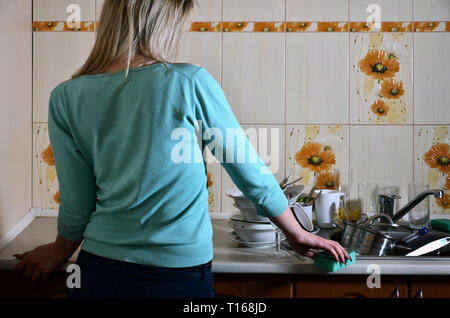 La ragazza, orinato e stanco di quotidiana mano-lavaggio piatti sorge accanto al bancone della cucina e un lavandino che è riempito con un rack di unwashed dishe Foto Stock