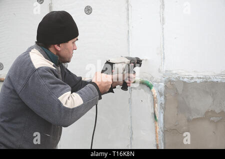 Un anziano operaio trapani un foro in una parete in polistirolo espanso per l'installazione successiva di un rinforzo di plastica il grano. Creazione di fori nel muro con un Foto Stock