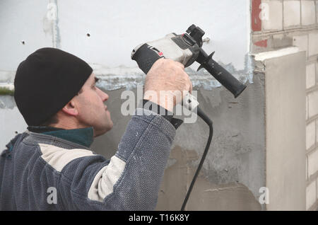 Un anziano operaio trapani un foro in una parete in polistirolo espanso per l'installazione successiva di un rinforzo di plastica il grano. Creazione di fori nel muro con un Foto Stock