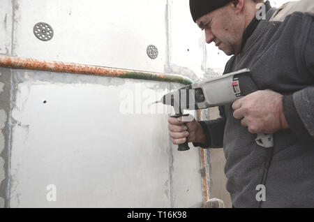 Un anziano operaio trapani un foro in una parete in polistirolo espanso per l'installazione successiva di un rinforzo di plastica il grano. Creazione di fori nel muro con un Foto Stock