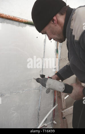 Un anziano operaio trapani un foro in una parete in polistirolo espanso per l'installazione successiva di un rinforzo di plastica il grano. Creazione di fori nel muro con un Foto Stock