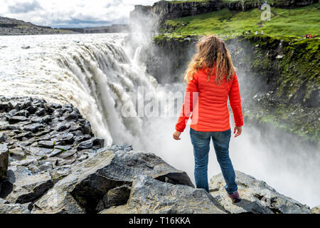 Giovane donna torna in piedi la visione di Dettifoss cascata su rocce che scorre acqua di irrorazione di nebbia giornata soleggiata in Islanda con camicia arancione e jeans Foto Stock