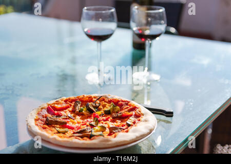 Pizza fresca sul tavolo di vetro terrazza fuori in Italia con salsa di pomodoro verdure e due bicchieri di vino rosso Bottiglia Foto Stock