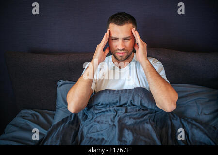 Concentrato giovane uomo tiene per mano da parte di testa. Egli tiene gli occhi chiusi. Ha mal di testa. Il ragazzo è sofferenza. Egli è coperto con una coperta Foto Stock