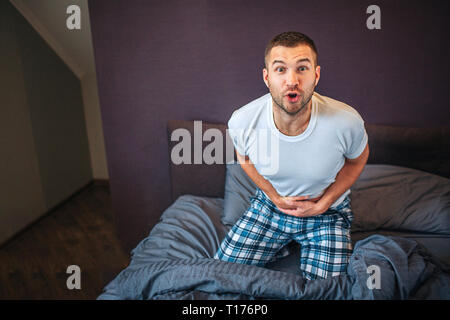 Si chiedeva il giovane uomo guarda sulla fotocamera con vista eccitato. Egli detiene hans su stomaco. Egli indossa pigiama. Guy vuole andare alla toilette. Foto Stock