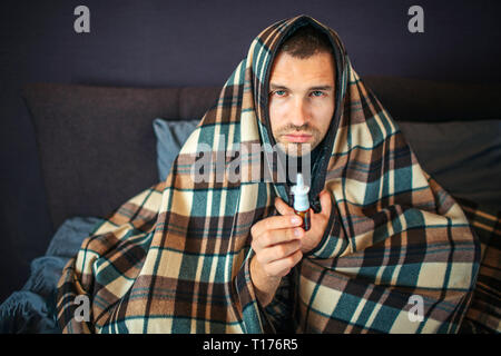 Malati giovane uomo guarda sulla fotocamera. Egli detiene il naso spray in mano. Il ragazzo è grave, egli è completamente coperto con una coperta. Giovane uomo è in camera da letto. Foto Stock