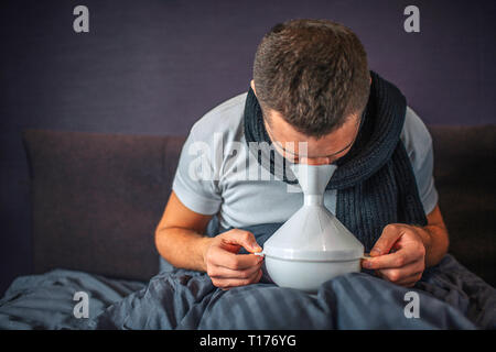 Immagine dell uomo usando un inalatore per il trattamento e per ottenere una migliore. È malato e malato. Guy detiene cosa bianca attenta e con entrambe le mani. Egli siede su Foto Stock