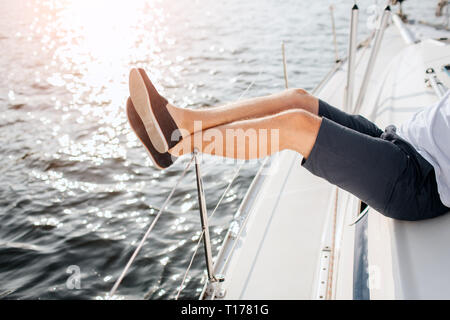 Vista di taglio del giovane di gambe che li trattiene fino sul bordo della scheda di yacht. Ragazzo indossa pantaloncini scuri, t-shirt bianco e nero scarpe. Egli è rilassato e tranquillo Foto Stock