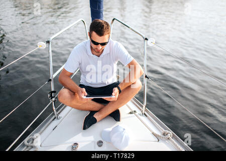 Giovane uomo si siede con le gambe incrociate e tenere premuto tablet in mani. Egli è calma e concentrata. Lo schermo è nero. Guy siede sulla prua dello yacht. Egli indossa Foto Stock