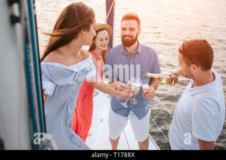 Bella foto di quattro persone in piedi insieme su yacht. Uomo in camicia bianca si riversa champaigne in bicchieri. Brunette guarda un altro uomo. Egli sorride Foto Stock