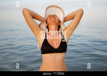 Donna di mezza età in un costume da bagno in mare.La donna in una spiaggia hat nell'oceano sollevato in alto le mani Foto Stock