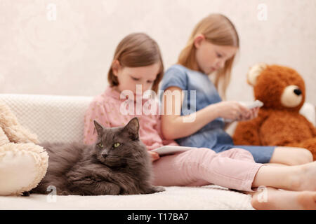 Due bambine (suore 7 e 8 anni) sta guardando un e-book e uno smartphone a casa sul divano con il loro gatto. Messa a fuoco selettiva. Foto Stock