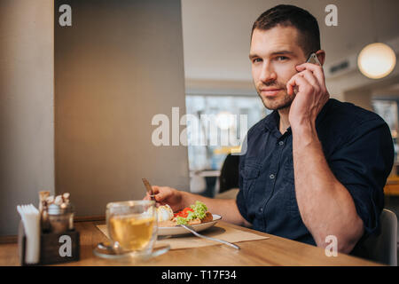 Il ragazzo è seduto in cafe e avente la cena. Anche lui sta parlando al telefono e guardando dritto alla telecamera. Egli sembra buono e fiducioso Foto Stock