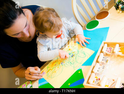 Vista ad angolo alto della madre che aiuta la giovane figlia ad esprimere il suo sé artistico su carta troppo colorata, seduto al tavolo da cucina. Foto Stock