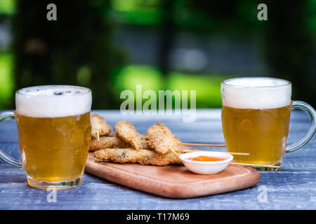 La birra snack set. Ali di pollo su bastoni e due boccali di birra. Sul tagliere e verde sullo sfondo di estate Foto Stock