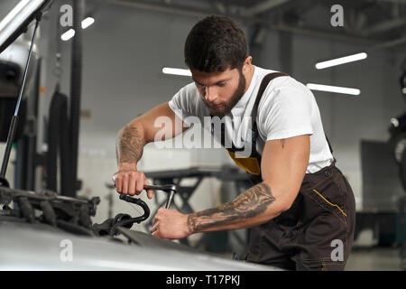 Auto riparazione e manutenzione. Giovani meccanica muscolare in tute e white t shirt riparazione auto in stazione autoservise. Bello barbuto lavoratore con i capelli scuri e tatuaggi sulle mani tenendo l'attrezzo. Foto Stock