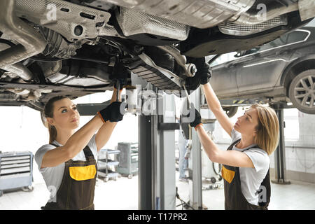 Due belle giovani donne in tute e guanti neri cercando, riparazione auto carro. Professional meccanica femminile concentrato sul fissaggio di automobile. Foto Stock