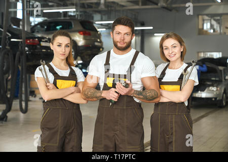 Tre meccanica professionale in piedi in autoservice, guardando la fotocamera. Belle Donne e brutale, uomo muscoloso con tatuaggi in posa insieme, tenendo delle chiavi dinamometriche e attrezzo. Foto Stock