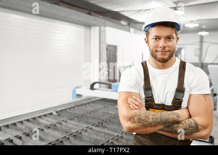 Ingegnere professionale e lavoratore di metallo in tute bianche, hardhat, stando in piedi in fabbrica. Bello, brutale uomo con tatuaggi in posa con mani incrociate, guardando la fotocamera e sorridente. Foto Stock