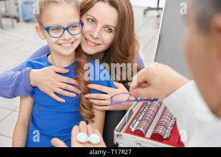 Pretty girl in vetri azzurrati e madre scegliendo occhio lenti per il trattamento e la correzione di visione in clinica medica. Medico oculista proponendo le lenti a contatto e occhiali. Foto Stock