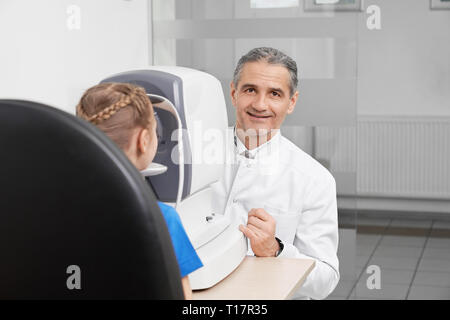 Dottore in medicina bianco rivestire l'esame della vista su attrezzature speciali in ottico ufficio di clinica medica. Oculista seduta a tavola con lampada a fessura, guardando la telecamera, sorridente. Foto Stock