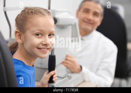 Paziente esame della vista ottico in ufficio. Bella ragazza adolescente in posa, sorridente e guardando la fotocamera. Oculista e lampada a fessura macchina su sfondo. Foto Stock