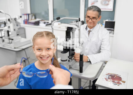 Bella ragazza sorridente e guardando la fotocamera. Mani femminili dando per scegliere i pazienti vetri blu. Medico all'ottico optometrista seduta a tavola con lampada a fessura in sala medica della clinica. Foto Stock