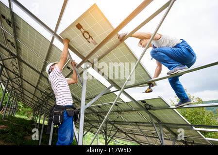 Installazione di impianti solari fotovoltaiche sistema del pannello. Due tecnici il sollevamento di un carico pesante modulo solare sulla piattaforma alta. Gli investimenti in energia alternativa, soldi s Foto Stock