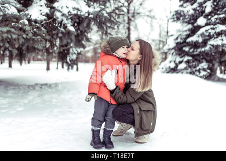 Una giovane madre baciare il suo bambino, ragazzo di 3-5 anni, in inverno nella foresta, contro uno sfondo derive di neve coperto di neve e gli alberi di Natale. Emozioni Foto Stock