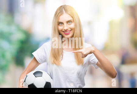 Giovane bella donna bionda tenendo palla calcio su sfondo isolato con sorpresa faccia puntare il dito a se stesso Foto Stock